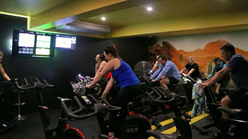 The spin room at CountryFit Gym in Mudgee is lively and energetic, featuring a group of participants engaged in a cycling class. Bright screens display workout metrics, while the vibrant decor adds to the motivating atmosphere.
