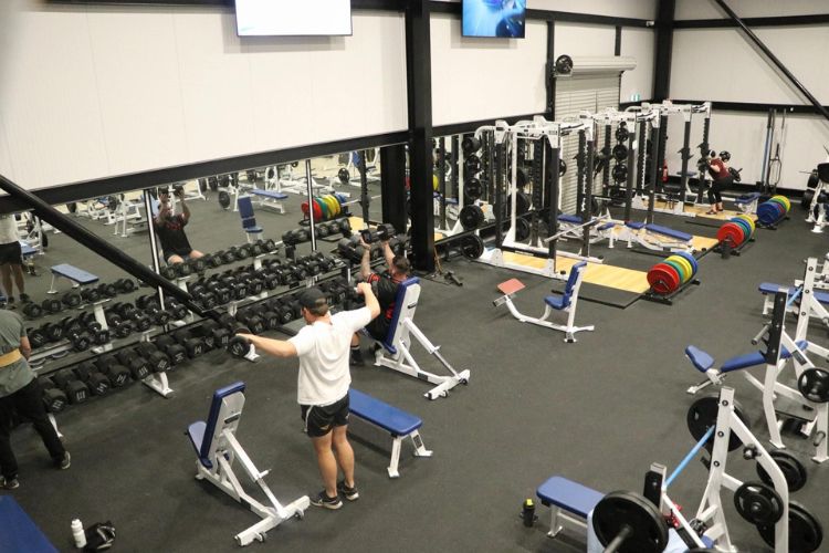 An interior view of CountryFit Gym in Mudgee, showcasing a well-equipped workout area. The image features various strength training equipment, including dumbbells and weight benches, with several gym-goers engaged in their workouts.