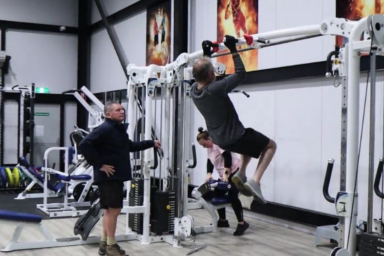 A scene inside CountryFit Gym in Mudgee, featuring a personal training session. A male trainer observes as a gym-goer performs pull-ups on a cable machine, demonstrating strength training.