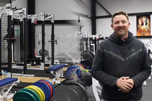 A split image showing CountryFit Gym Mudgee environment on the left with various weightlifting equipment and benches, while on the right, a man stands smiling, wearing a black Nike hoodie. He appears engaged and approachable, reflecting a positive attitude towards fitness and well-being. The background includes a vibrant artwork, enhancing the gym's atmosphere.