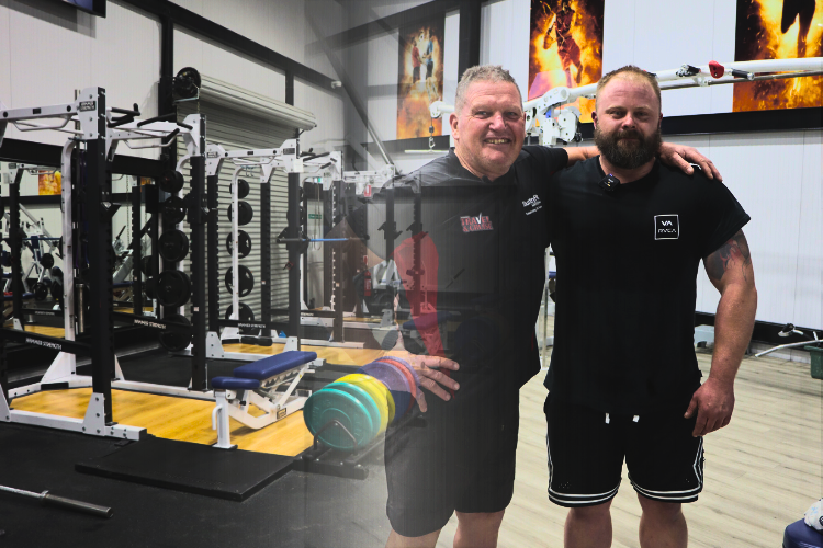 Two men stand together in a well-equipped gym, smiling and posing for the camera. Glen, on the left, wears a black shirt from CountryFit and showcases a friendly demeanour, while Scott, on the right, in a black t-shirt with a logo, displays a proud expression. The background features various fitness equipment, highlighting the supportive fitness community at CountryFit Gym in Mudgee.
