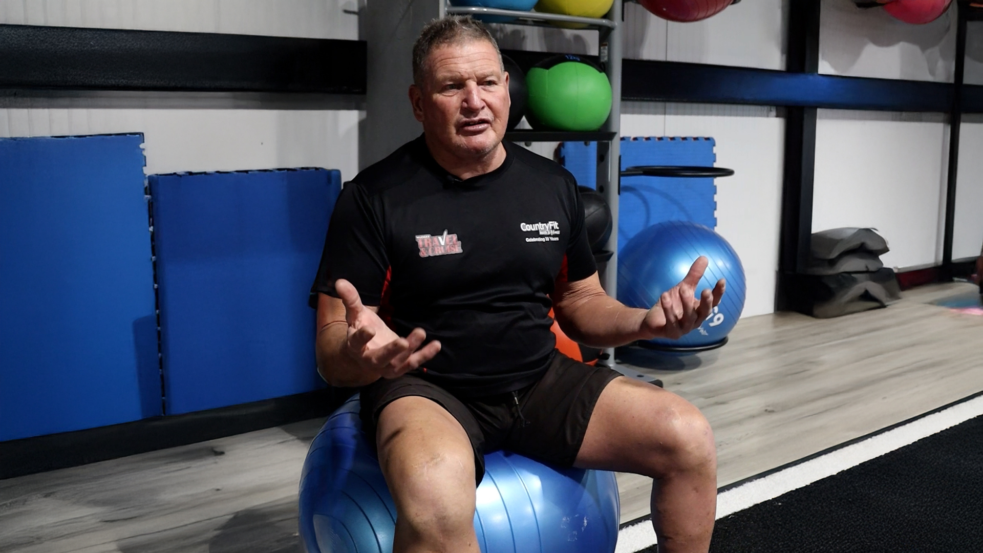 Glenn in a black CountryFit shirt sits in front of a lively gym scene where women are engaged in a boxing workout. The background showcases various fitness equipment, emphasising a dynamic and supportive fitness community. Glenn radiates positivity, reflecting the welcoming atmosphere of CountryFit Gym in Mudgee.