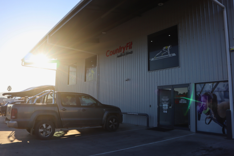 The exterior of CountryFit Gym in Mudgee, NSW, showcasing a modern metal building with large windows. The CountryFit logo is displayed on the upper side of the building, and a pickup truck is parked in front. Sunlight is shining through, highlighting the welcoming entrance with a promotional image of a woman exercising. This conveys the inclusive and accessible fitness environment that CountryFit offers for all fitness levels.